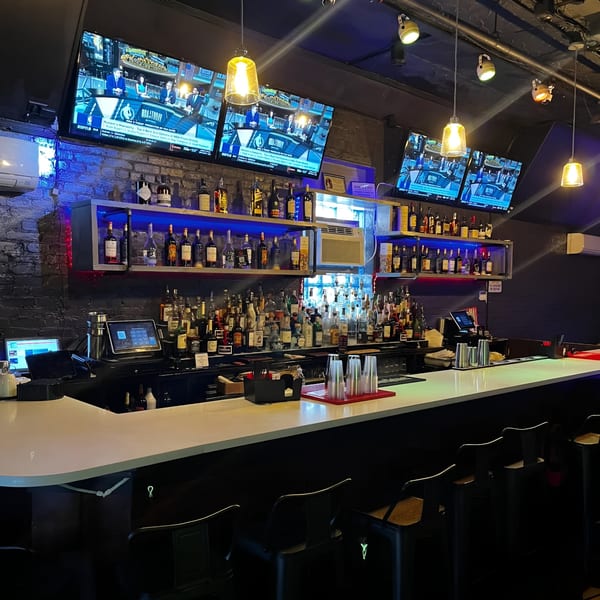A well-stocked bar with multiple liquor bottles, bar stools, and TVs showing sports, illuminated by blue and yellow lights.