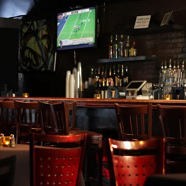 A dimly lit bar area with a TV showing football, bottles on shelves, and wooden bar stools.