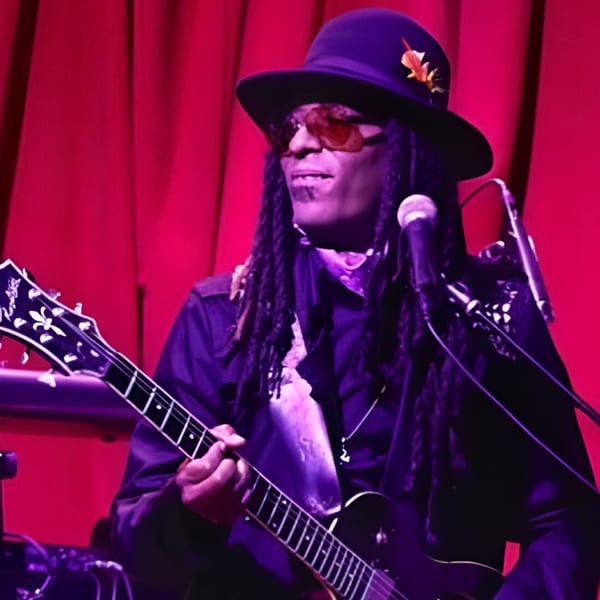 Musician with dreadlocks, hat, and sunglasses playing an electric guitar on a red-lit stage.