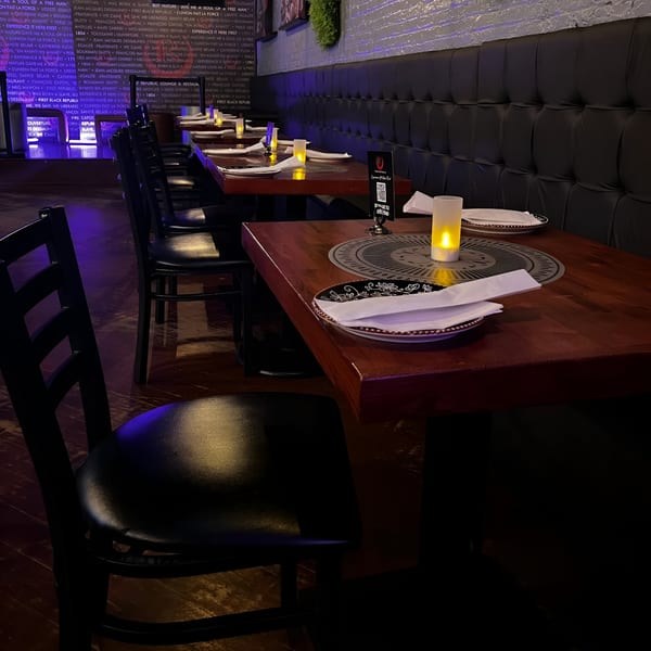Dimly lit dining area with dark tables, chairs, and a tufted bench, featuring glowing candles and wall text.