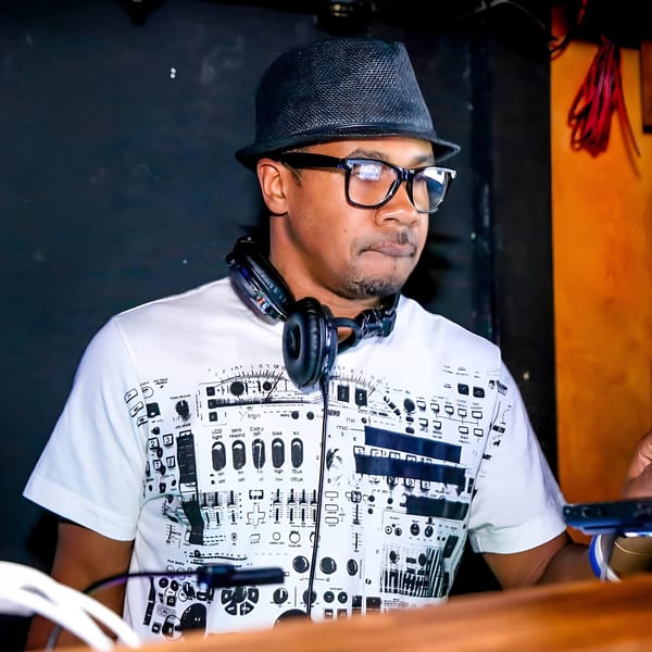 A DJ wearing a fedora, glasses, and headphones around his neck, playing music at First Republic.